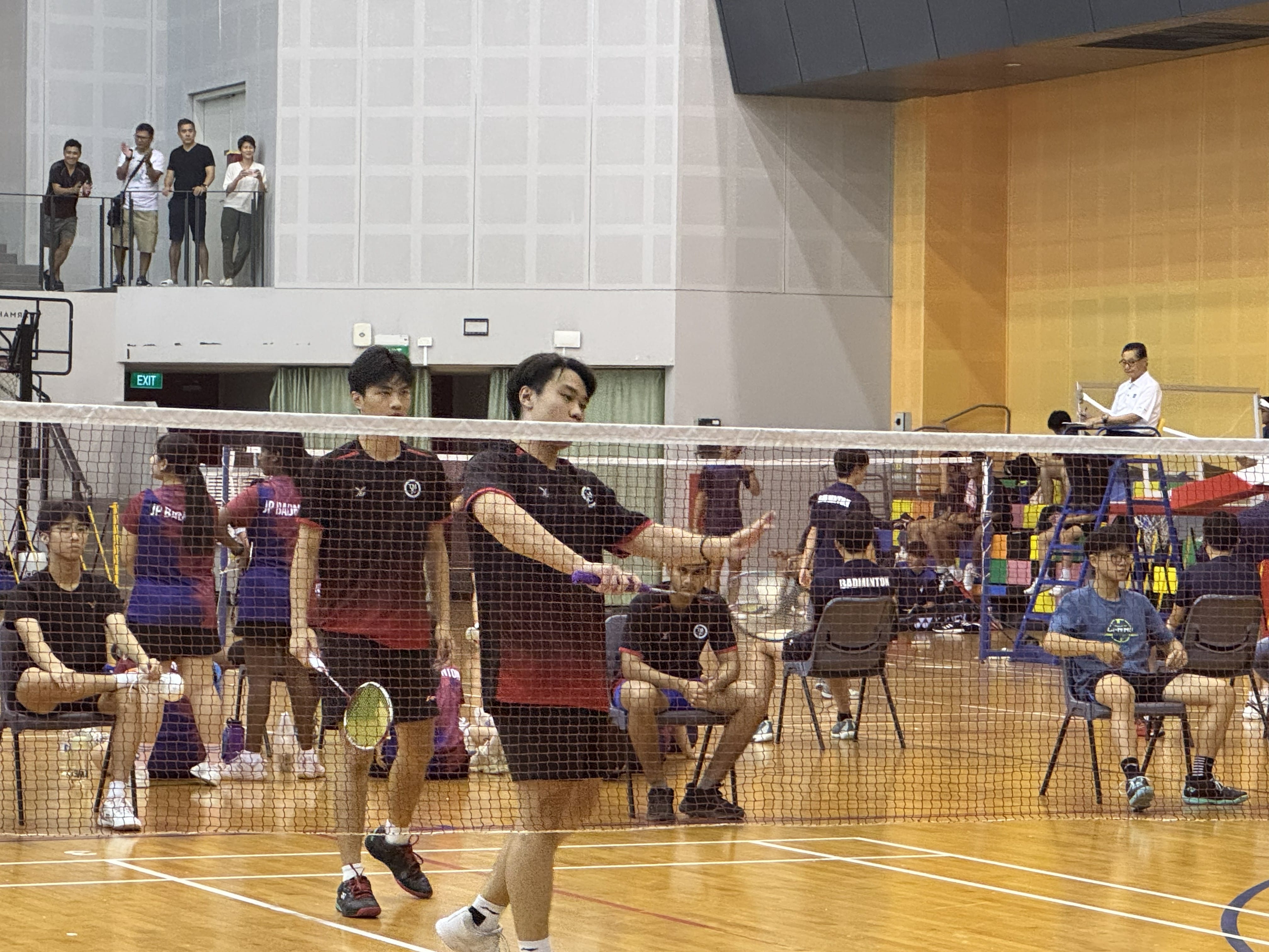 Two TMJC boys in dark jerseys compete in a doubles badminton match at the National School Games, with spectators watching from an upper level gallery.